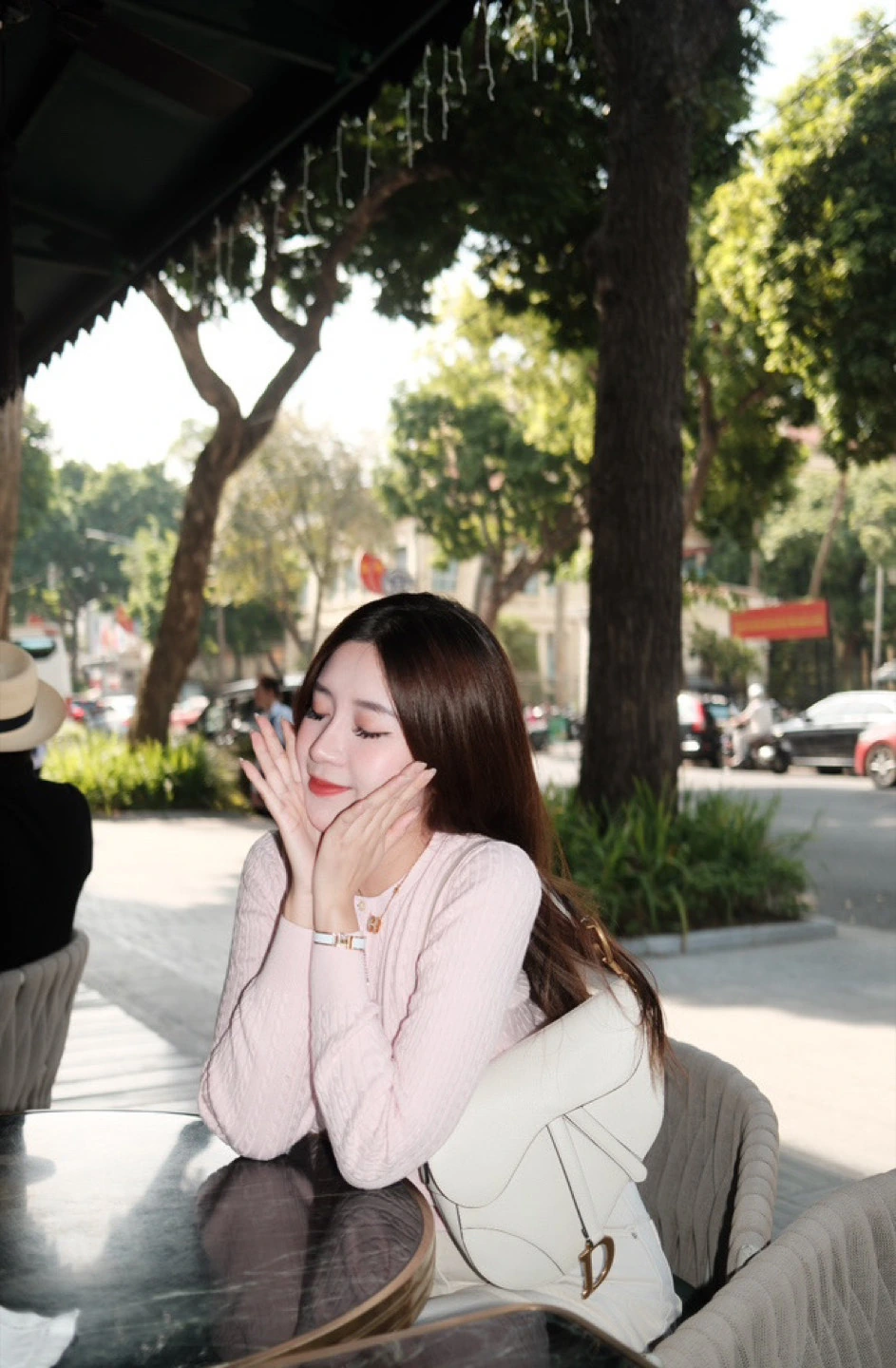 A woman in a pale pink top resting her face in her hands at an outdoor cafe table.