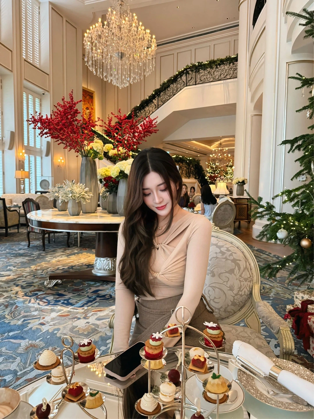 A woman seated at a table of small cakes beneath a chandelier in an ornate dining room.