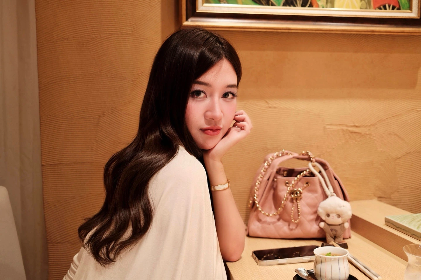 A woman looking back at the camera from a restaurant counter, beside a pink handbag and a small cup.