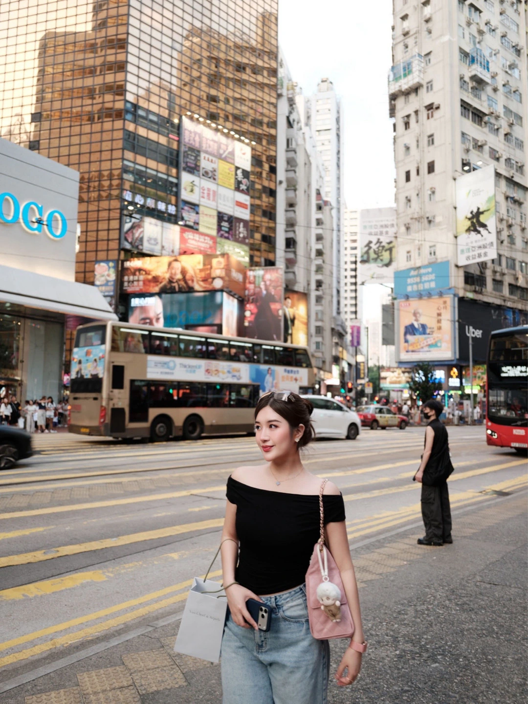 A woman in a black off-shoulder top and jeans standing near a busy city crossing.