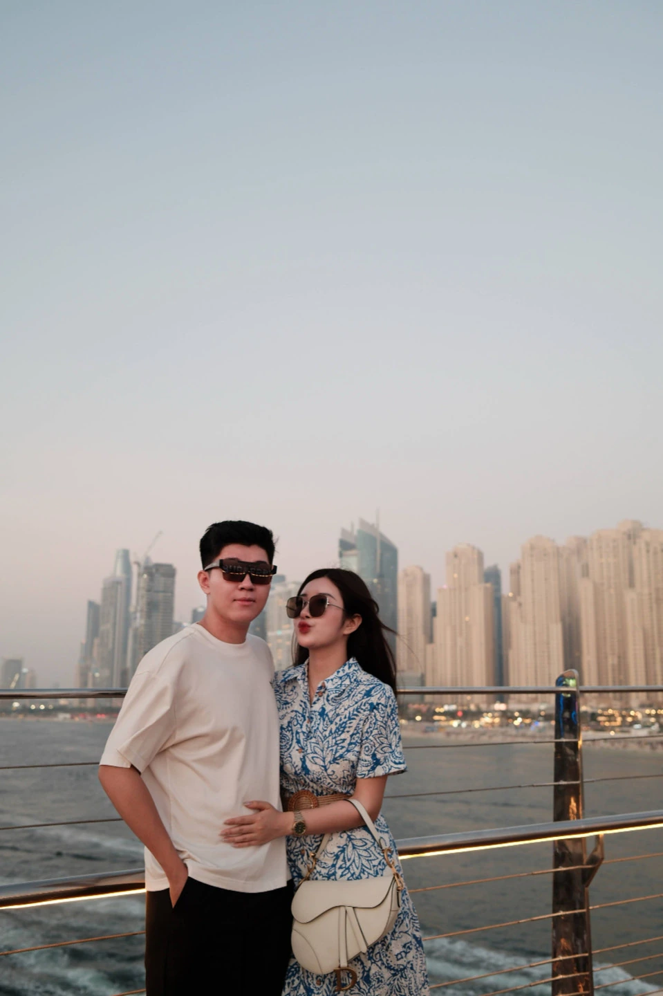 The couple standing beside a waterfront railing with a city skyline behind them.