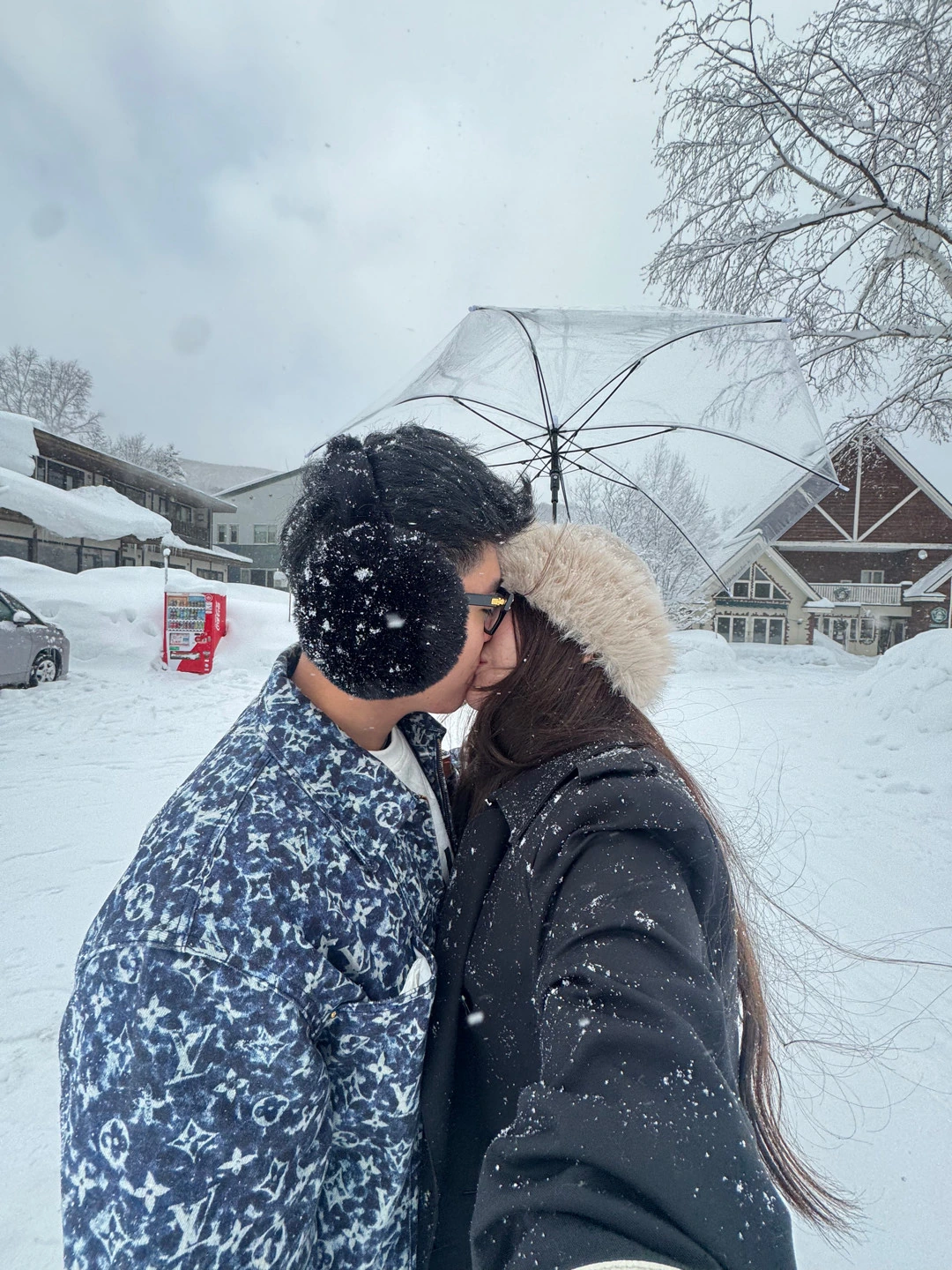 The couple kissing under a clear umbrella as snow falls around them.