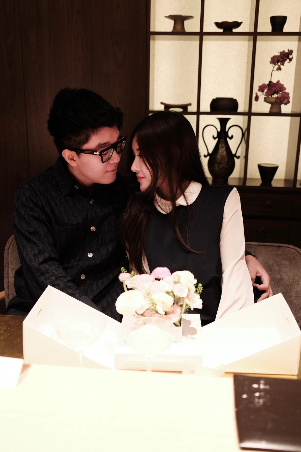 The couple looking at each other across a table with a small bouquet of flowers.
