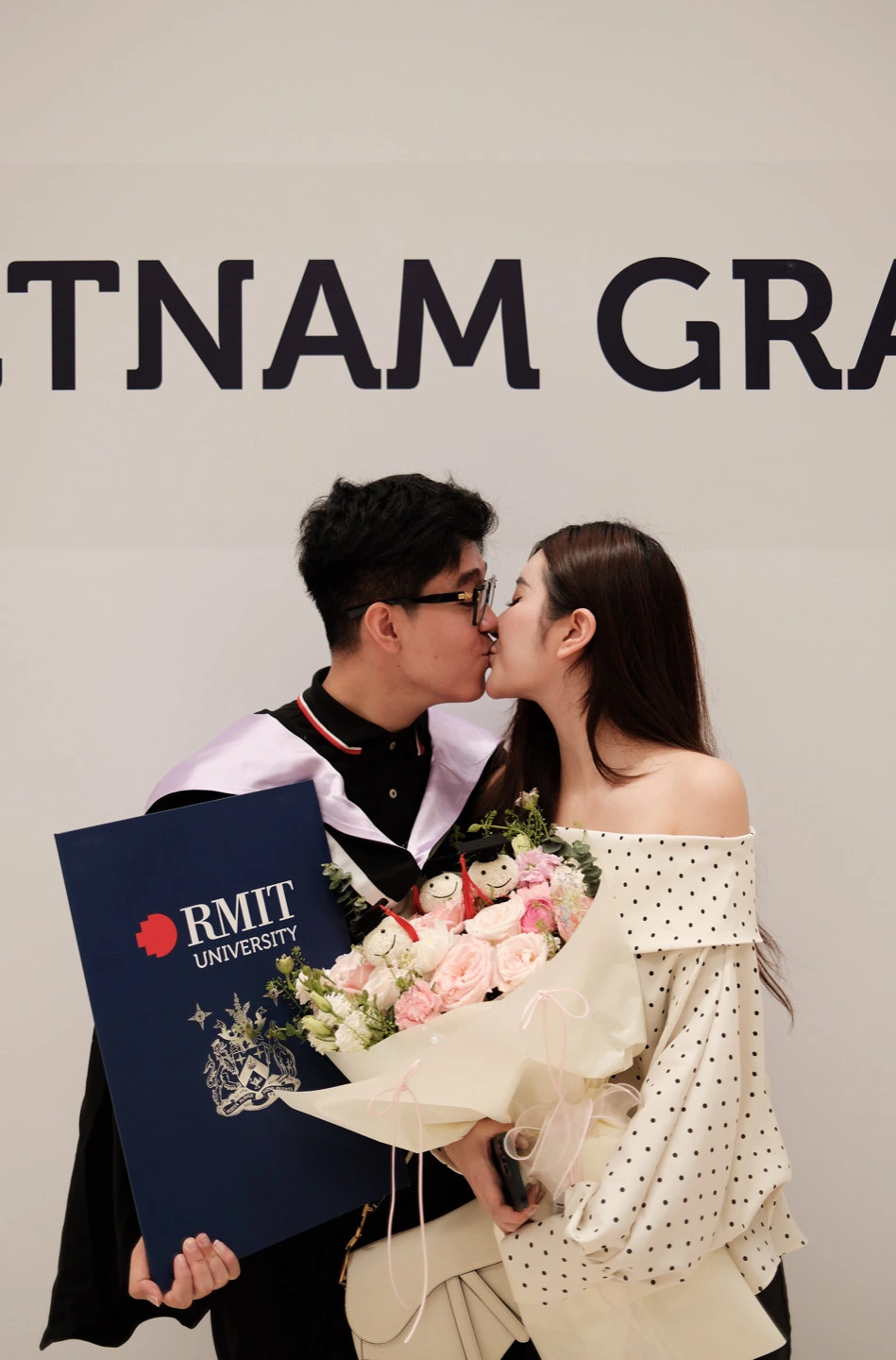 The couple sharing a kiss while holding flowers and a graduation folder.